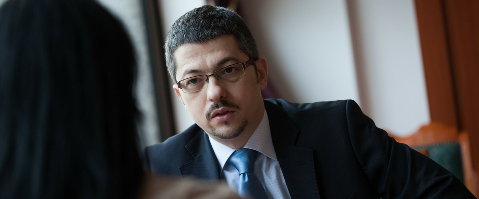 A man in a suit sits at a desk during an interview